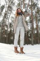 A young and joyful Caucasian girl in a brown coat holds a snowball in a snow-covered forest in winter. Fisheye Photo