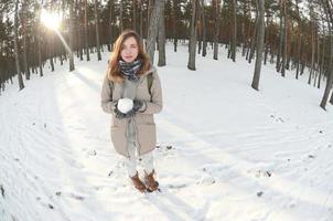 A young and joyful Caucasian girl in a brown coat holds a snowball in a snow-covered forest in winter. Fisheye Photo