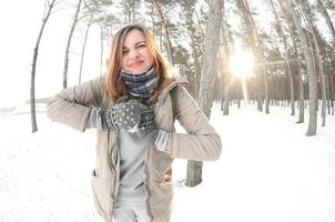 A young and joyful Caucasian girl in a brown coat sculpts a snowball in a snow-covered forest in winter. Games with snow in the open air. Fisheye Photo