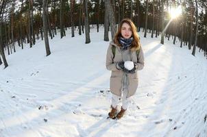 una joven y alegre chica caucásica con un abrigo marrón sostiene una bola de nieve en un bosque cubierto de nieve en invierno. foto de ojo de pez