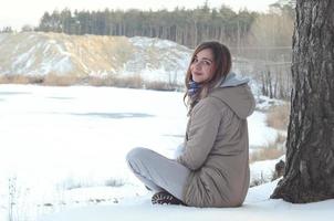 A young Caucasian girl in a brown coat is sitting near a cliff in the background of a horizon line between the sky and a frozen lake in winter time photo