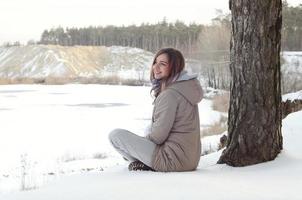 A young Caucasian girl in a brown coat is sitting near a cliff in the background of a horizon line between the sky and a frozen lake in winter time photo