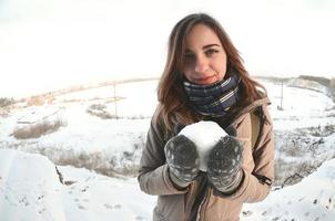 una joven y alegre chica caucásica con un abrigo marrón sostiene una bola de nieve frente a una línea de horizonte entre el cielo y un lago congelado en invierno. foto de ojo de pez