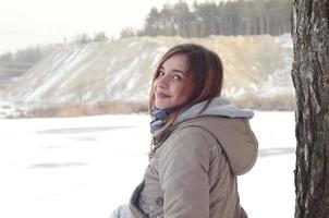 A young Caucasian girl in a brown coat is sitting near a cliff in the background of a horizon line between the sky and a frozen lake in winter time photo