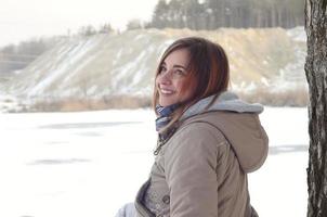 A young Caucasian girl in a brown coat is sitting near a cliff in the background of a horizon line between the sky and a frozen lake in winter time photo