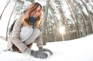 A young and joyful Caucasian girl in a brown coat sculpts a snowball in a snow-covered forest in winter. Games with snow in the open air. Fisheye Photo