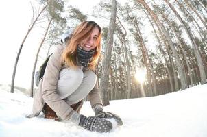 una joven y alegre chica caucásica con un abrigo marrón esculpe una bola de nieve en un bosque cubierto de nieve en invierno. juegos con nieve al aire libre. foto de ojo de pez