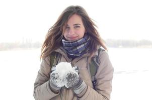 A young and joyful Caucasian girl in a brown coat holds a snowball in the background of a horizon line between the sky and a frozen lake in winter photo