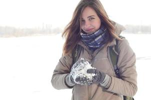 A young and joyful Caucasian girl in a brown coat holds a snowball in the background of a horizon line between the sky and a frozen lake in winter photo