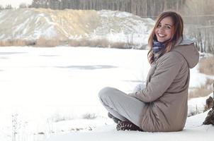 A young Caucasian girl in a brown coat is sitting near a cliff in the background of a horizon line between the sky and a frozen lake in winter time photo