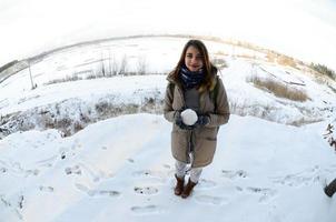 una joven y alegre chica caucásica con un abrigo marrón sostiene una bola de nieve frente a una línea de horizonte entre el cielo y un lago congelado en invierno. foto de ojo de pez