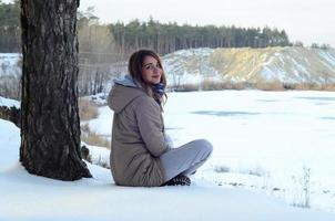 A young Caucasian girl in a brown coat is sitting near a cliff in the background of a horizon line between the sky and a frozen lake in winter time photo