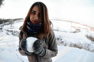 A young and joyful Caucasian girl in a brown coat holds a snowball in front of a horizon line between the sky and a frozen lake in winter. Fisheye Photo