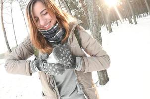 A young and joyful Caucasian girl in a brown coat sculpts a snowball in a snow-covered forest in winter. Games with snow in the open air. Fisheye Photo