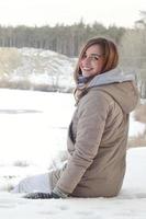 A young Caucasian girl in a brown coat is sitting near a cliff in the background of a horizon line between the sky and a frozen lake in winter time photo