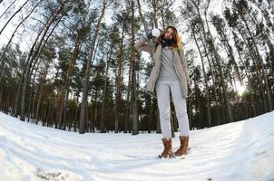 una joven y alegre chica caucásica con un abrigo marrón sostiene una bola de nieve en un bosque cubierto de nieve en invierno. foto de ojo de pez