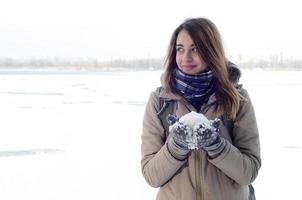 A young and joyful Caucasian girl in a brown coat holds a snowball in the background of a horizon line between the sky and a frozen lake in winter photo