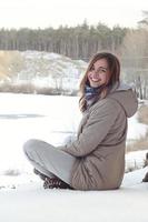 A young Caucasian girl in a brown coat is sitting near a cliff in the background of a horizon line between the sky and a frozen lake in winter time photo