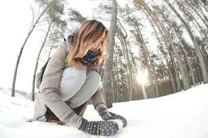 A young and joyful Caucasian girl in a brown coat sculpts a snowball in a snow-covered forest in winter. Games with snow in the open air. Fisheye Photo