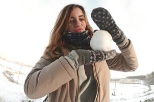 A young and joyful Caucasian girl in a brown coat holds a snowball in front of a horizon line between the sky and a frozen lake in winter. Fisheye Photo