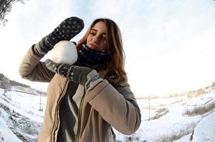 una joven y alegre chica caucásica con un abrigo marrón sostiene una bola de nieve frente a una línea de horizonte entre el cielo y un lago congelado en invierno. foto de ojo de pez