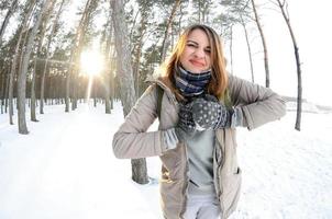 A young and joyful Caucasian girl in a brown coat sculpts a snowball in a snow-covered forest in winter. Games with snow in the open air. Fisheye Photo