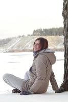 A young Caucasian girl in a brown coat is sitting near a cliff in the background of a horizon line between the sky and a frozen lake in winter time photo