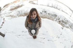 una joven y alegre chica caucásica con un abrigo marrón sostiene una bola de nieve frente a una línea de horizonte entre el cielo y un lago congelado en invierno. foto de ojo de pez