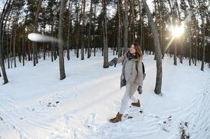 A young and joyful Caucasian girl in a brown coat is throwing a snowball in a snow-covered forest in winter. Games with snow in the open air. Fisheye Photo