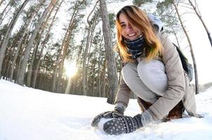 A young and joyful Caucasian girl in a brown coat sculpts a snowball in a snow-covered forest in winter. Games with snow in the open air. Fisheye Photo