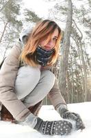 A young and joyful Caucasian girl in a brown coat sculpts a snowball in a snow-covered forest in winter. Games with snow in the open air. Fisheye Photo