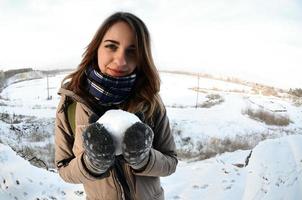 A young and joyful Caucasian girl in a brown coat holds a snowball in front of a horizon line between the sky and a frozen lake in winter. Fisheye Photo