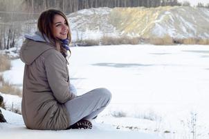 A young Caucasian girl in a brown coat is sitting near a cliff in the background of a horizon line between the sky and a frozen lake in winter time photo