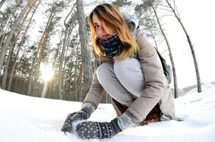 A young and joyful Caucasian girl in a brown coat sculpts a snowball in a snow-covered forest in winter. Games with snow in the open air. Fisheye Photo