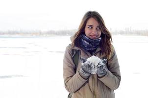 A young and joyful Caucasian girl in a brown coat holds a snowball in the background of a horizon line between the sky and a frozen lake in winter photo