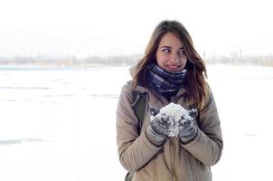 una joven y alegre chica caucásica con un abrigo marrón sostiene una bola de nieve en el fondo de una línea de horizonte entre el cielo y un lago congelado en invierno foto