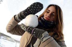 una joven y alegre chica caucásica con un abrigo marrón sostiene una bola de nieve frente a una línea de horizonte entre el cielo y un lago congelado en invierno. foto de ojo de pez