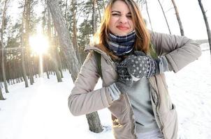 A young and joyful Caucasian girl in a brown coat sculpts a snowball in a snow-covered forest in winter. Games with snow in the open air. Fisheye Photo