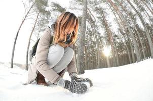 A young and joyful Caucasian girl in a brown coat sculpts a snowball in a snow-covered forest in winter. Games with snow in the open air. Fisheye Photo