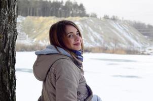 A young Caucasian girl in a brown coat is sitting near a cliff in the background of a horizon line between the sky and a frozen lake in winter time photo