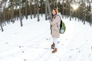 A young and joyful Caucasian girl in a brown coat holds a snowball in a snow-covered forest in winter. Fisheye Photo
