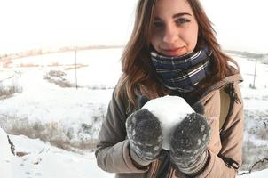 A young and joyful Caucasian girl in a brown coat holds a snowball in front of a horizon line between the sky and a frozen lake in winter. Fisheye Photo