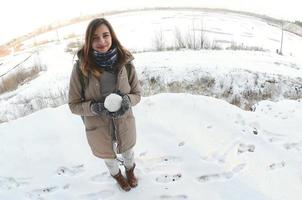 una joven y alegre chica caucásica con un abrigo marrón sostiene una bola de nieve frente a una línea de horizonte entre el cielo y un lago congelado en invierno. foto de ojo de pez