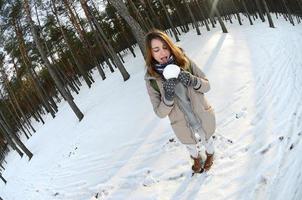 A young and joyful Caucasian girl in a brown coat holds a snowball in a snow-covered forest in winter. Fisheye Photo