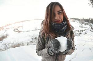 una joven y alegre chica caucásica con un abrigo marrón sostiene una bola de nieve frente a una línea de horizonte entre el cielo y un lago congelado en invierno. foto de ojo de pez