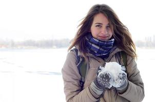 A young and joyful Caucasian girl in a brown coat holds a snowball in the background of a horizon line between the sky and a frozen lake in winter photo