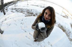 una joven y alegre chica caucásica con un abrigo marrón sostiene una bola de nieve frente a una línea de horizonte entre el cielo y un lago congelado en invierno. foto de ojo de pez