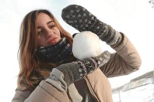 A young and joyful Caucasian girl in a brown coat holds a snowball in front of a horizon line between the sky and a frozen lake in winter. Fisheye Photo