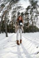 una joven y alegre chica caucásica con un abrigo marrón sostiene una bola de nieve en un bosque cubierto de nieve en invierno. foto de ojo de pez