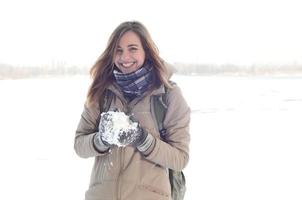 A young and joyful Caucasian girl in a brown coat holds a snowball in the background of a horizon line between the sky and a frozen lake in winter photo